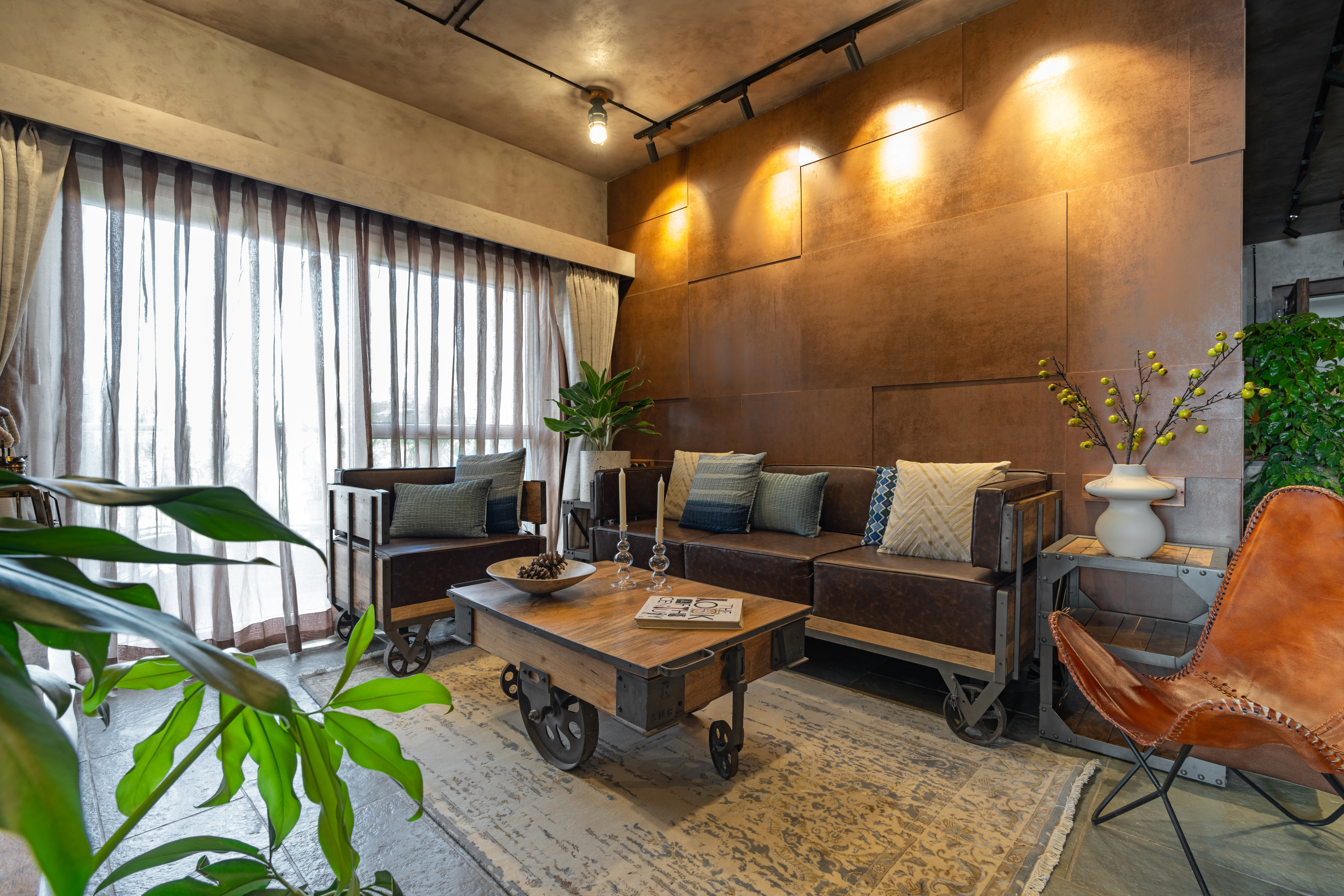 Living room with a patinated copper-panel wall: leather sofa on cart wheels with blue and ivory cushions, a timber cart coffee table holding a bowl and candlesticks, a pale faded rug over stone flooring, a tan leather butterfly chair and a white vase of yellow berry branches.